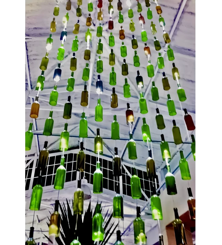 The installation consists of a series of green-tinted glass bottles suspended by wires, with strategic lighting, 
forming a kind of waterfall with a large vertical extension. The bottles, arranged at different heights, create 
an unusual and dramatic visual effect. At the bottom, bottles in a rectangular box simulate a “waterfall,” 
causing an explosion of lights, shadows, and emotions.