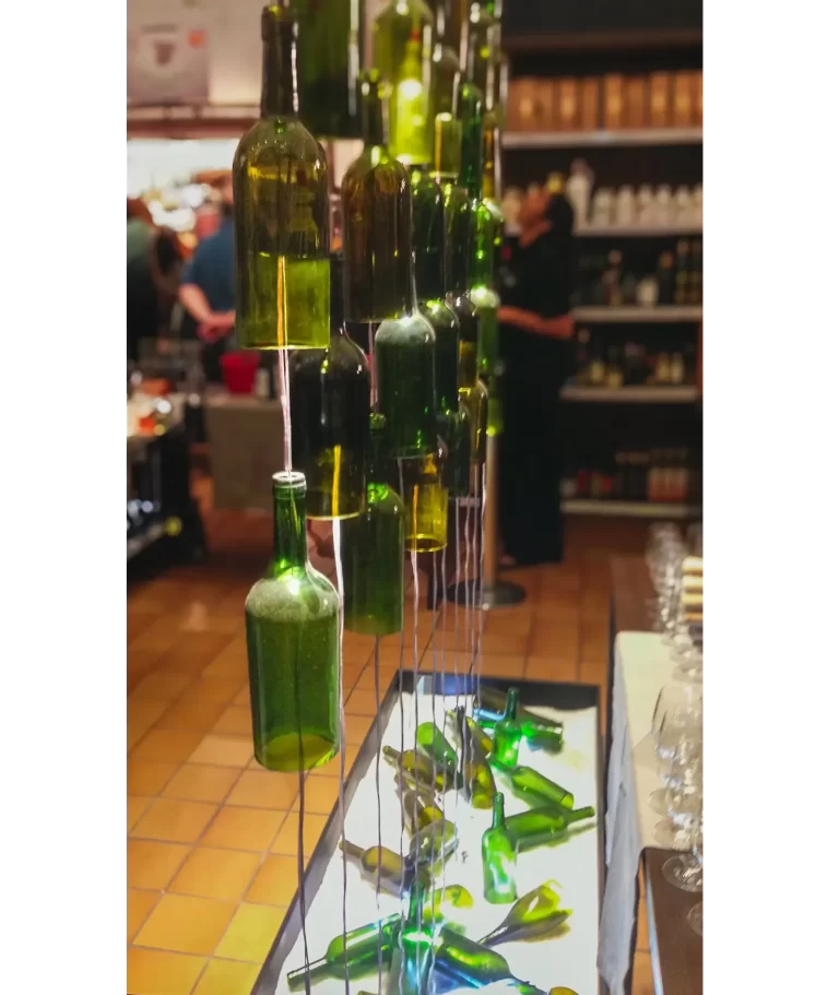 The installation consists of a series of green-tinted glass bottles suspended by wires, with strategic lighting, 
forming a kind of waterfall with a large vertical extension. The bottles, arranged at different heights, create 
an unusual and dramatic visual effect. At the bottom, bottles in a rectangular box simulate a “waterfall,” 
causing an explosion of lights, shadows, and emotions.